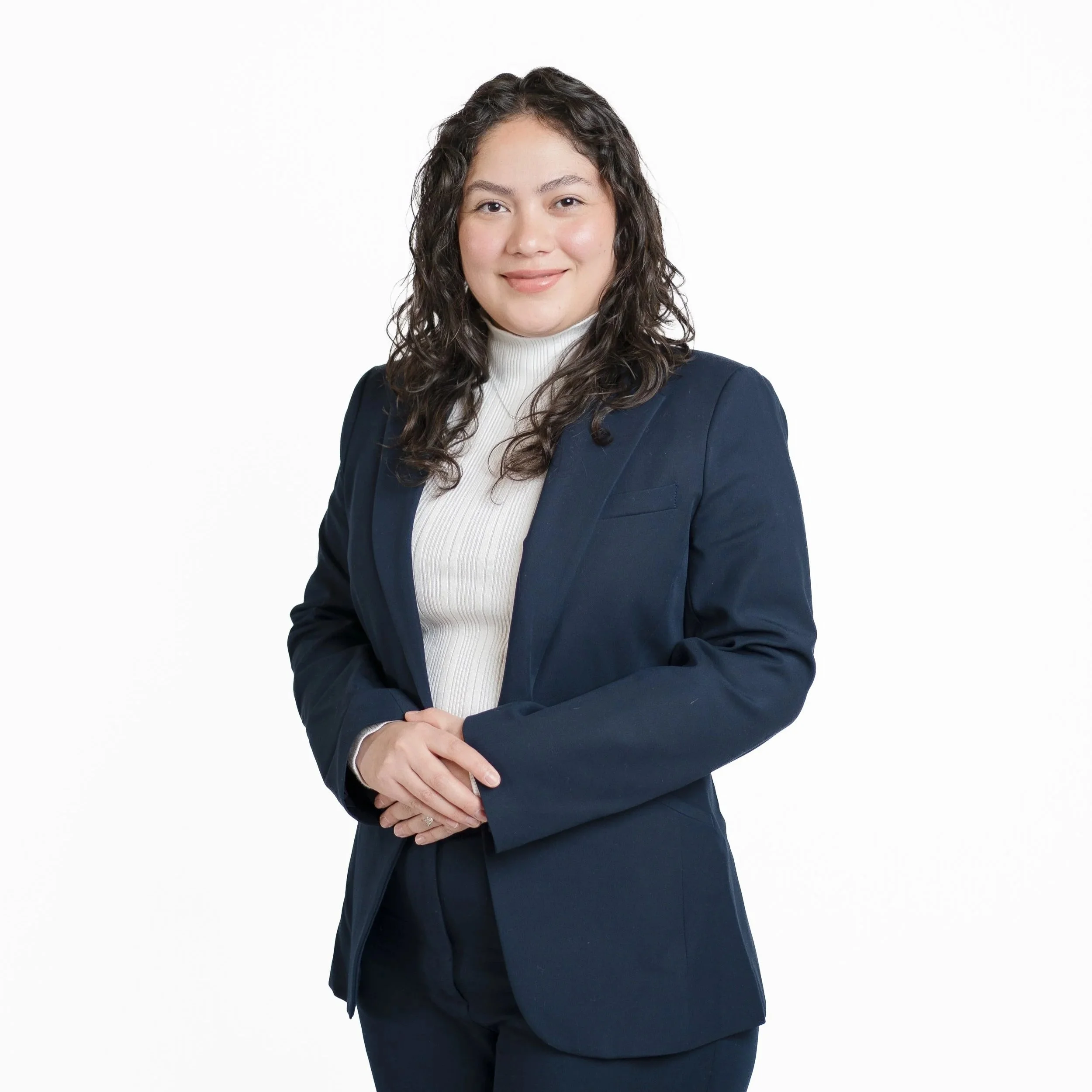 A woman with curly dark hair wearing a white turtleneck and navy blazer, smiling confidently against a plain white background.
