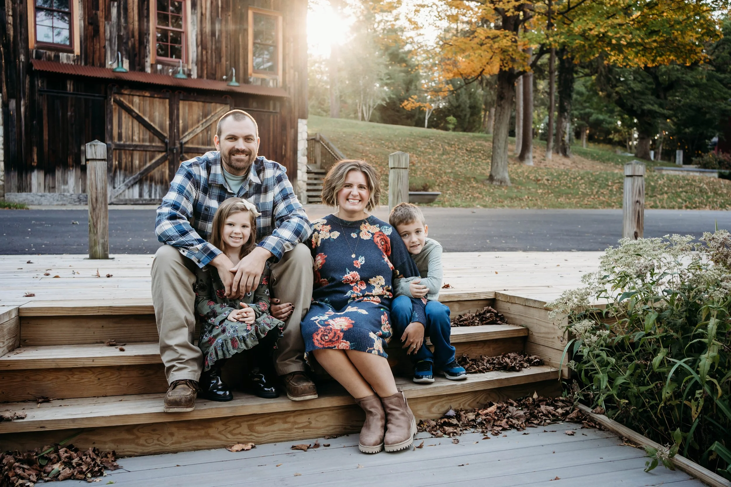 A family of four sitting on wooden steps outdoors during fall, with a rustic wooden building and trees with autumn leaves in the background.