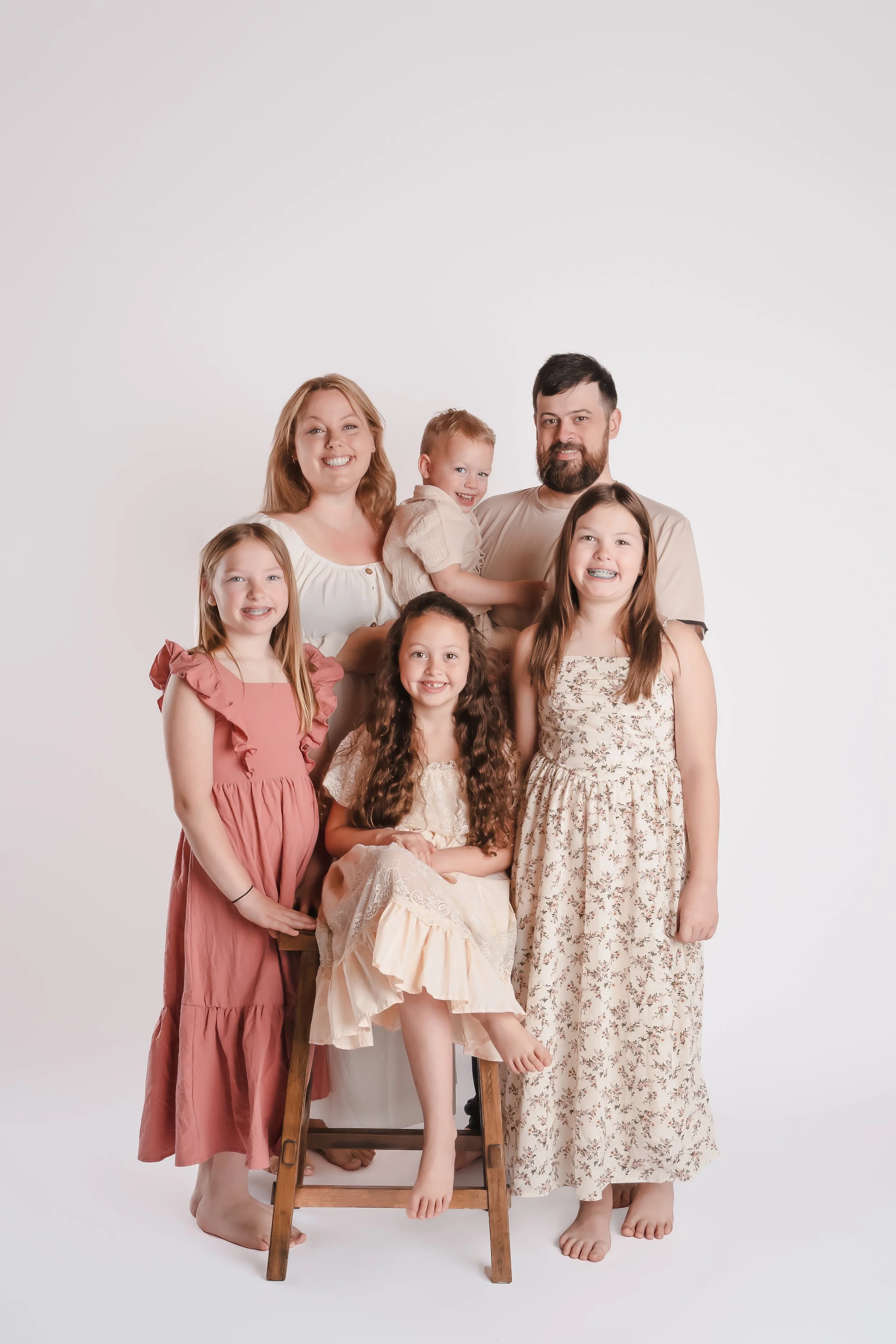 A multigenerational family of seven posing together in a studio with a plain white background. They are all smiling and wearing light-colored, casual, and floral clothing.