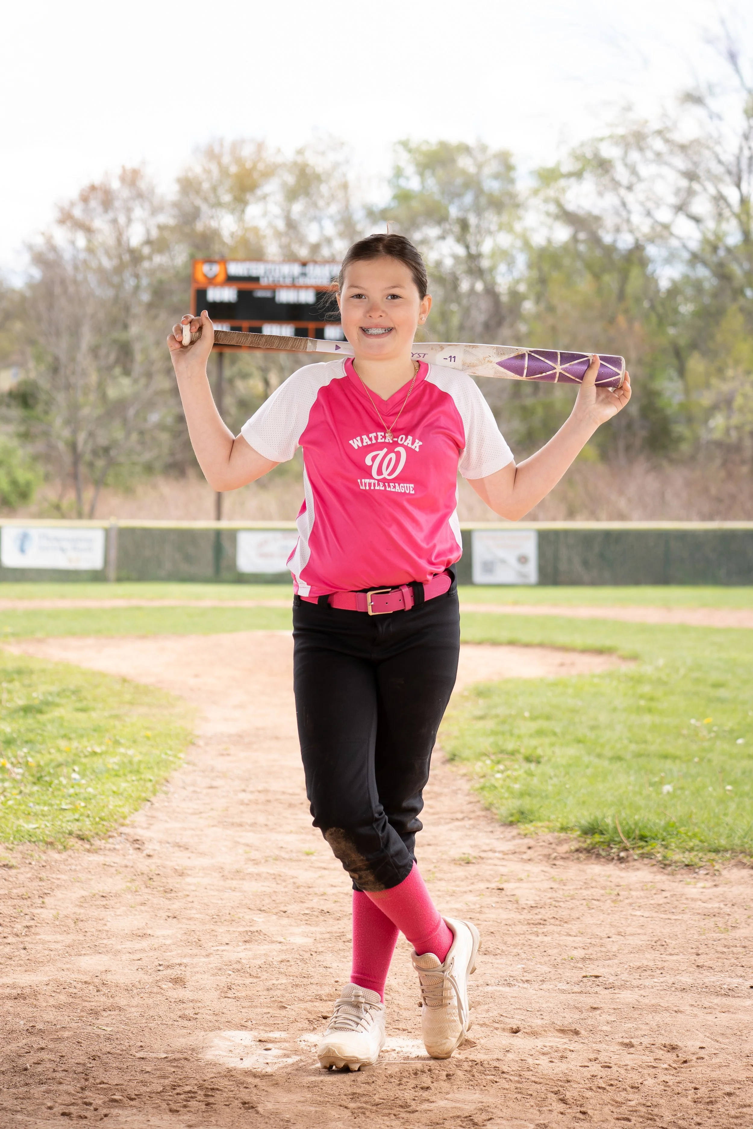 A young girl wearing a pink softball jersey, black pants, and pink socks is standing on a baseball diamond with a bat resting on her shoulders. She is smiling and outdoors on a baseball field.