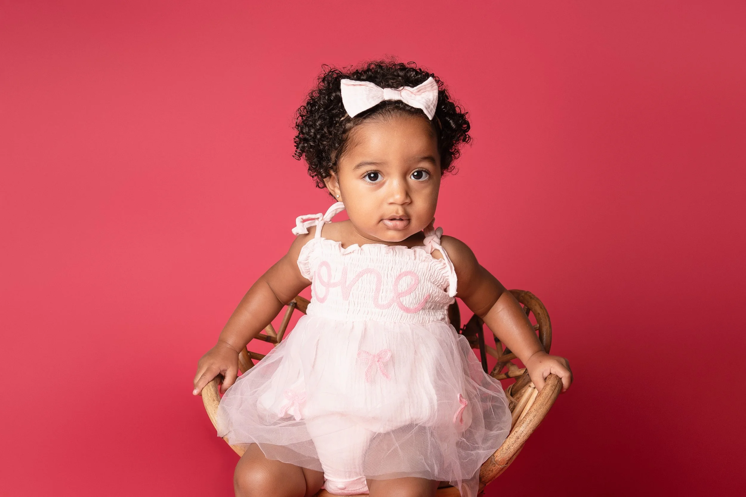 A young girl with curly hair and a pink bow, wearing a light pink dress, sitting on a wicker chair against a pink background.