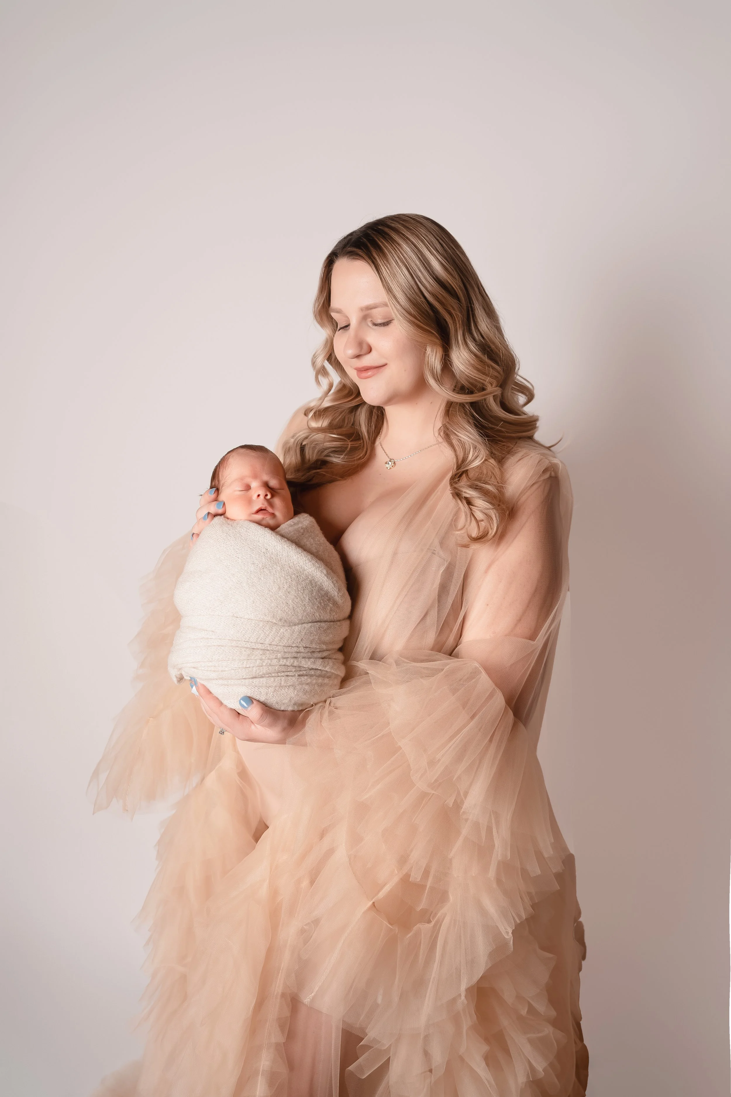 A woman with long curly blonde hair holding a sleeping newborn wrapped in a beige blanket. She is wearing a beige tulle gown and a silver necklace, set against a plain off-white background.