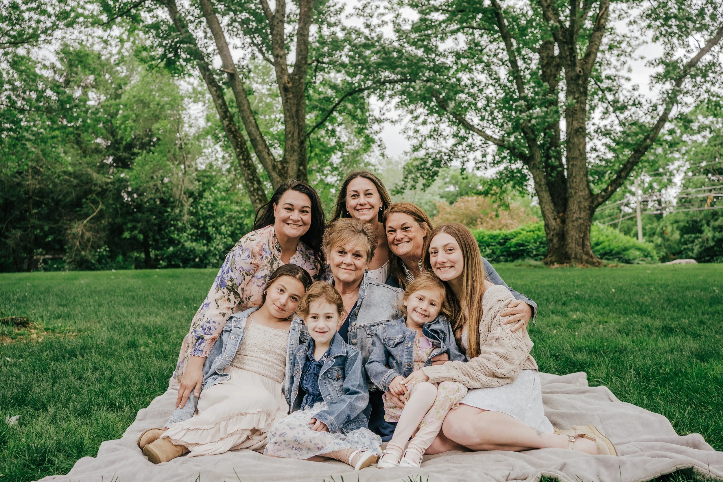 A group of women and children sitting and lying on a blanket on grass in a park, with trees and greenery in the background.