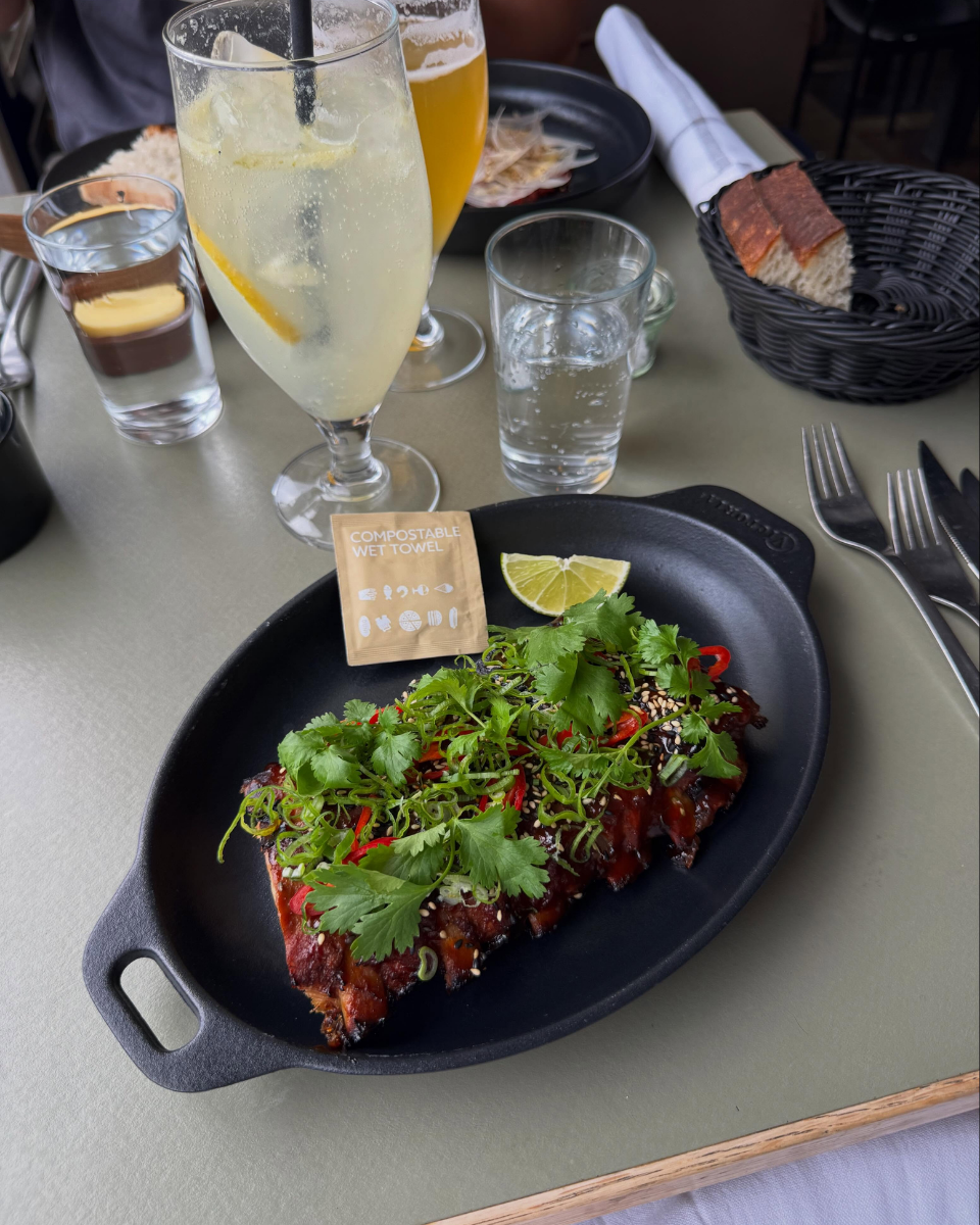En tallerken med BBQ-ribs toppet med frisk koriander, forårsager et limekryds ved siden af, omgivet af drikkevarer, herunder sodavand, øl og en lemonad. Der er også et stykke brød i en sort kurv, og bestik ligger på tallerkenen, alt på et spisebord.