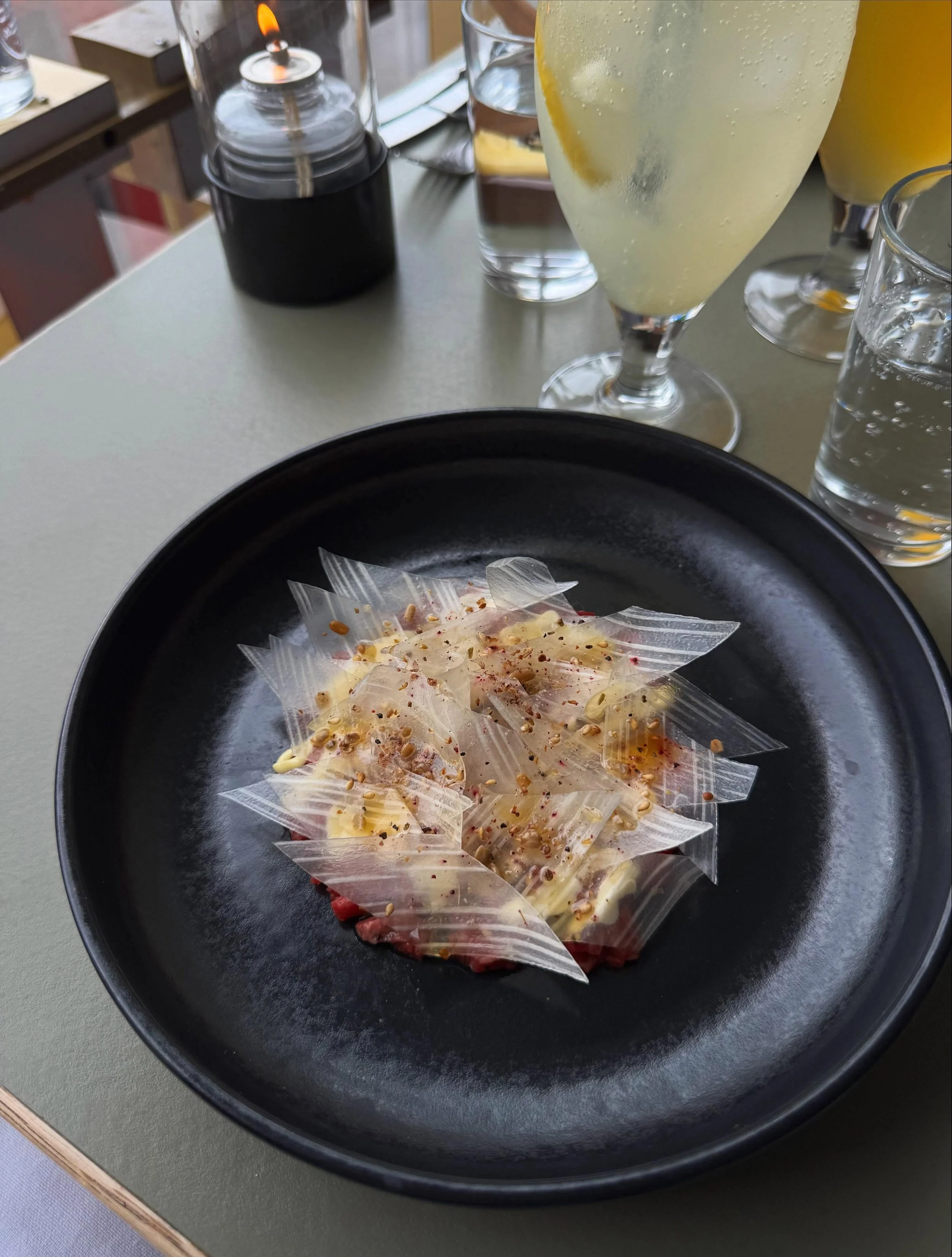 Billede af en sort tallerken med frosne druer dækket af hvid chokolade og drysset med hakkede nødder. Der er flere glas med vand og en champagneskål med mousserende vin med citron i baggrunden.