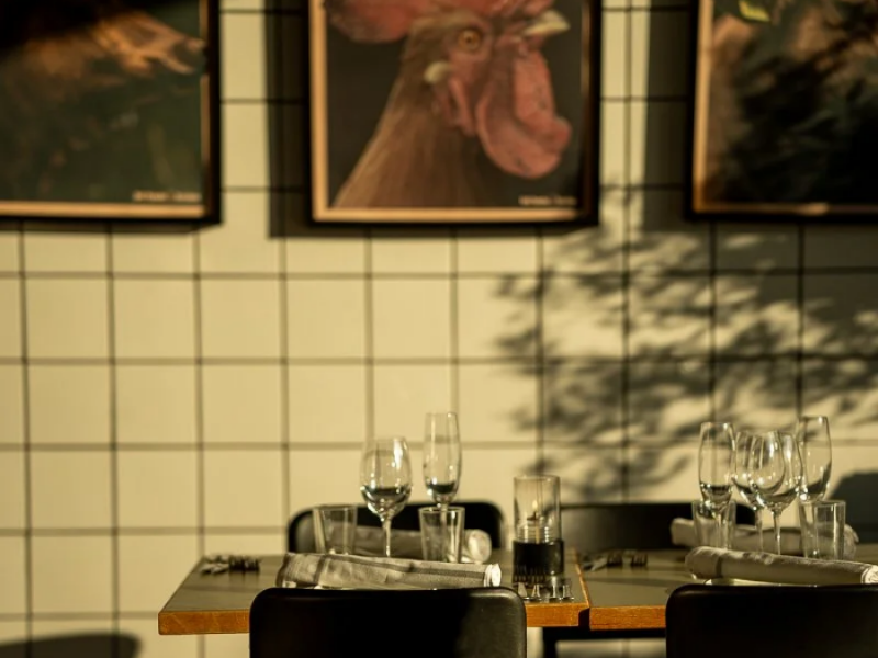 En restaurantbord er sat op med glas, bestik og servietter, med billeder af en høne på væggen i baggrunden.