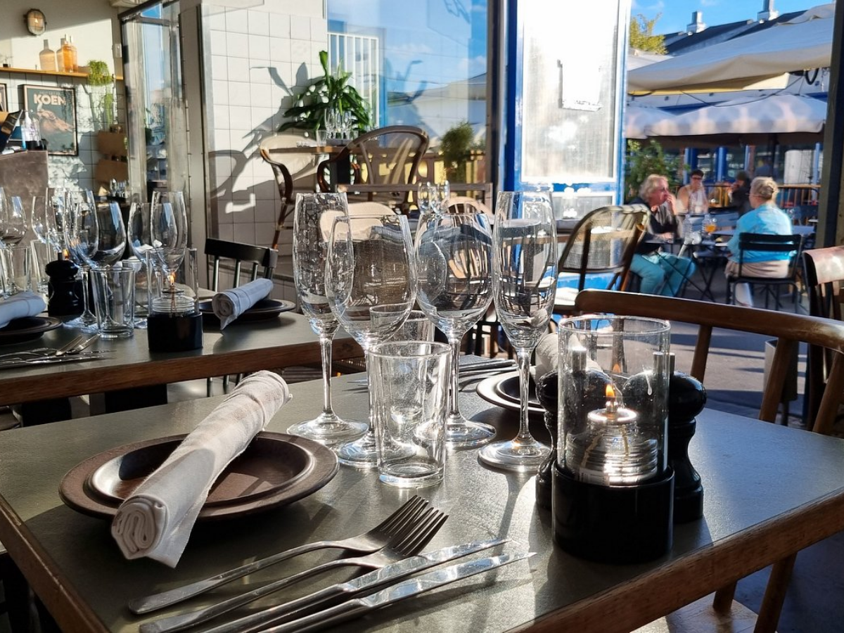 Indretning af en restaurant eller café med dækket bord, flere glas, bestik, tallerkener og stearinlys på et bord i baggrunden.