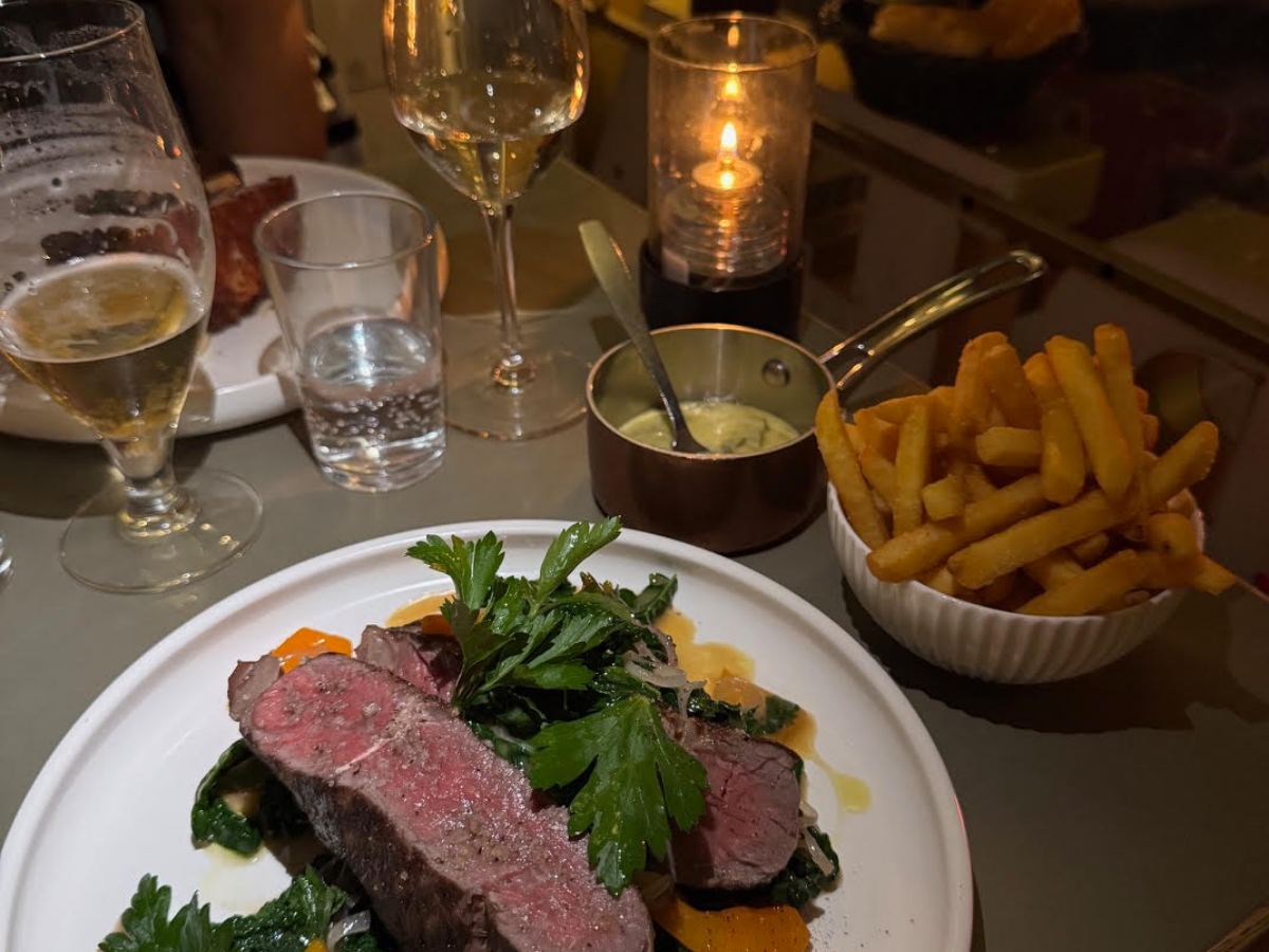 Et minutstykke oksekød med urter og grøntsager på en tallerken, omgivet af champagneglas, et glas vand, en skål med pommes frites, en lille gryde med sovs og et stearinlys i en lysestage.