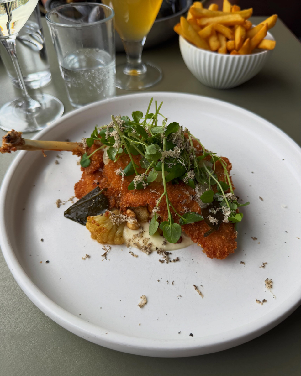En tallerken med paneret kød, toppet med grønne spirer og revet ost, serveret på et hvidt fad. I baggrunden er der en skål med pommes frites, flere glas med drikkevarer, herunder et øl og en guava drink.