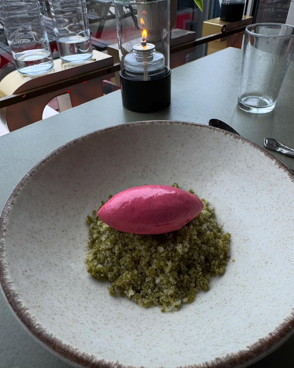 Billedet viser en tallerken med en grøn crumble og en stor, lyserød skumkantet skefuld på toppen, sandsynligvis en dessert. På bordet er der også et tomt glas og en drink- eller stearinlys-lampe i baggrunden.
