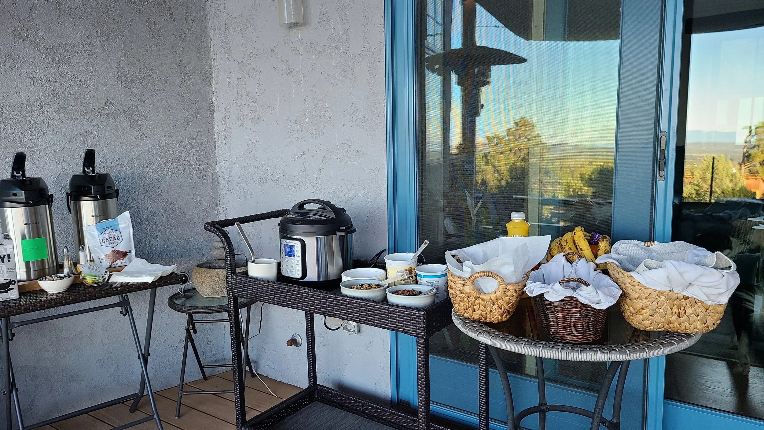 Outdoor breakfast setup with coffee dispensers, instant pot, condiments, and baskets of bananas and bread on a patio.