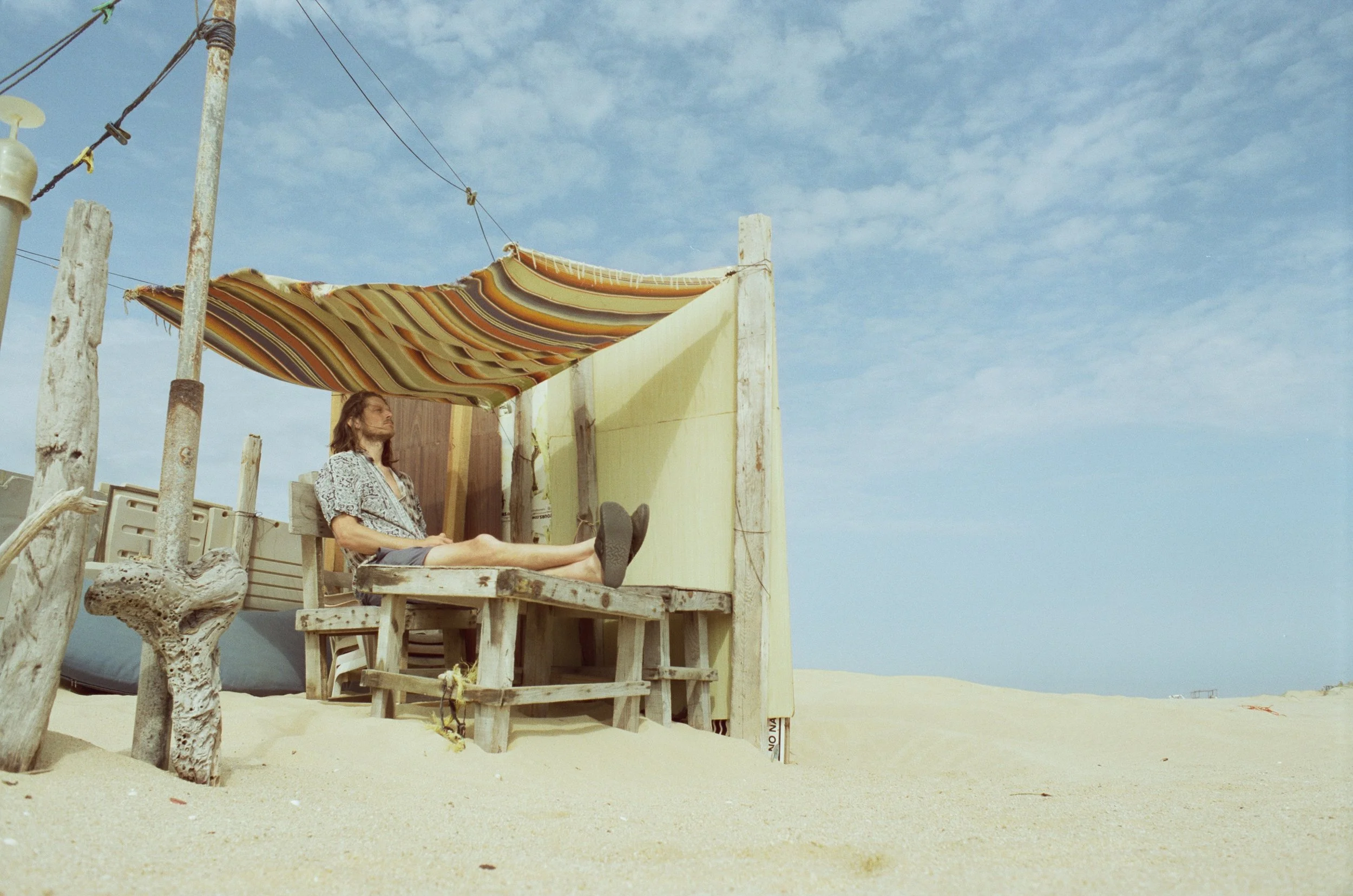 A person with long hair relaxing on a wooden beach hut with a striped canopy, set on a sandy beach under a partly cloudy sky.