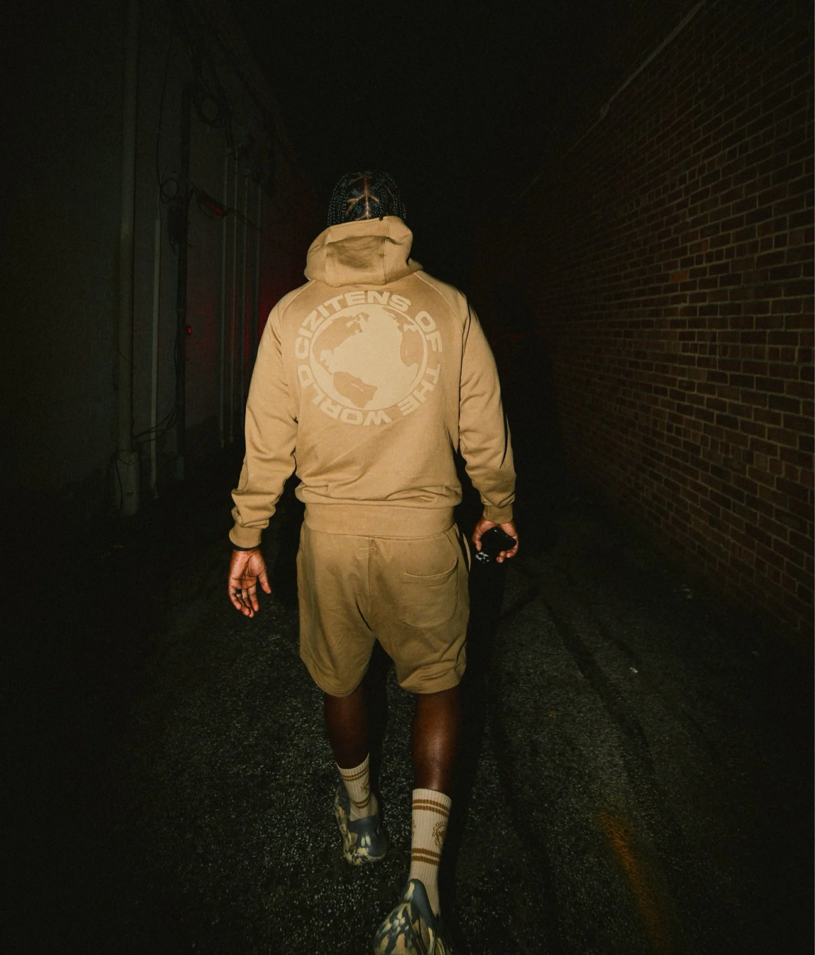 A person walking down a dark alley at night, wearing a beige hoodie with a globe graphic and text on the back, beige shorts, patterned socks, and sneakers.
