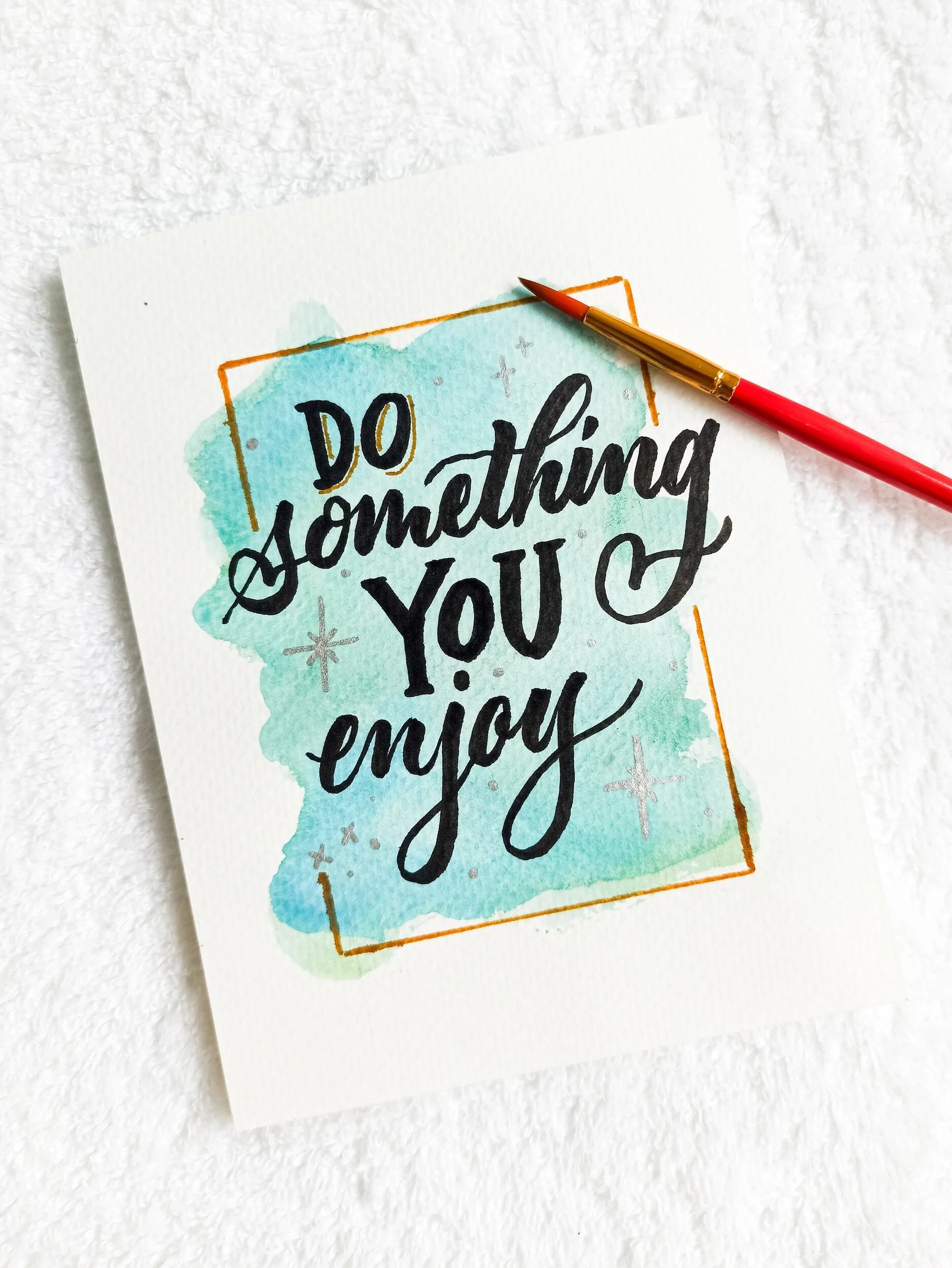 Wenskaart met de tekst 'Do something you enjoy', handgeschreven in zwart op een blauwe achtergrond, met een rood penseel ernaast op een witte ondergrond.