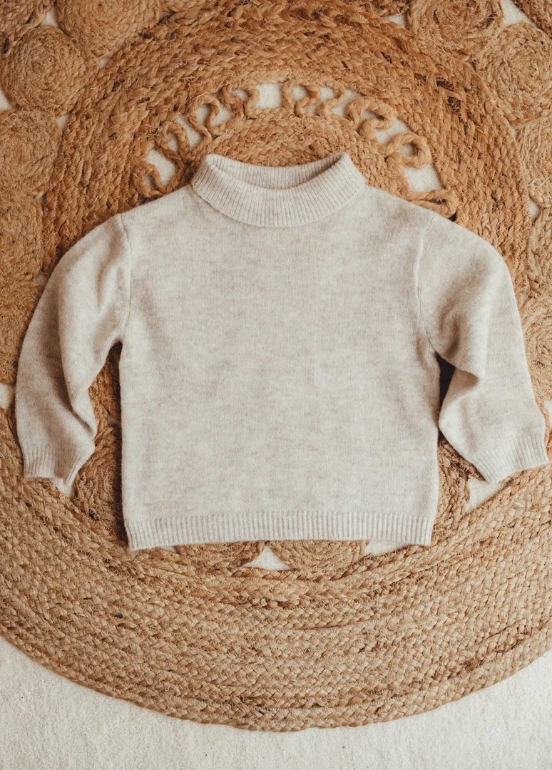 A small, cream-colored sweater with a turtleneck laid flat on a round, woven, tan-colored rug.