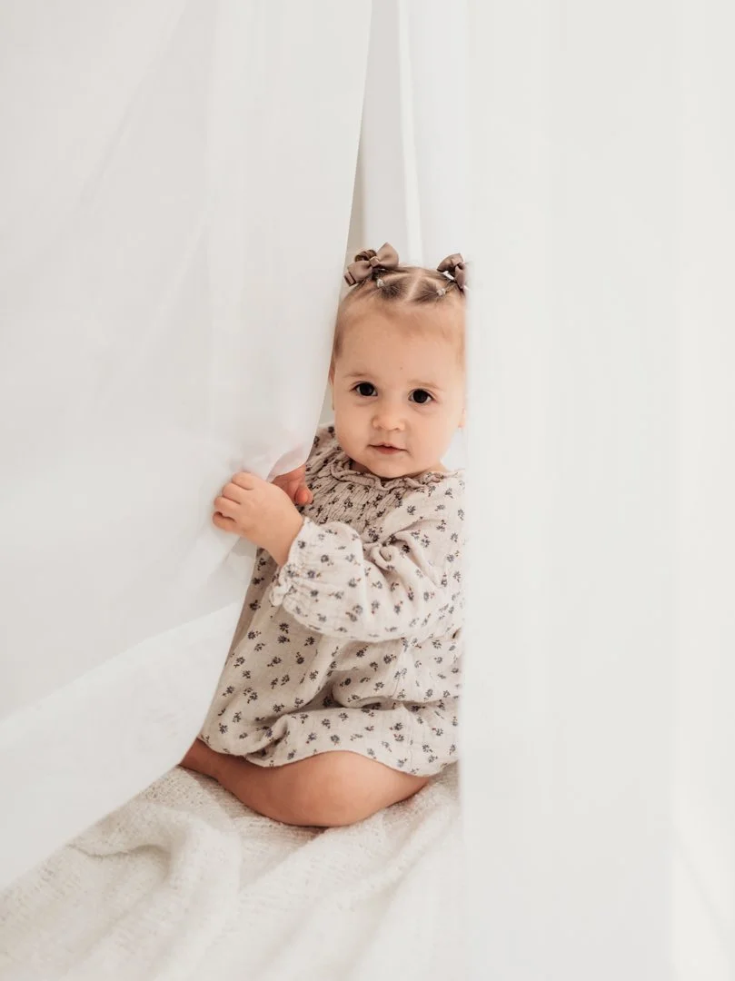 A young girl with light hair sitting on a soft, white blanket while peeking out from behind a white curtain or bedspread.