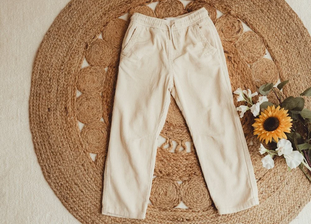 A pair of cream-colored corduroy pants placed on a circular woven rug with a sunflower and white flowers beside them.