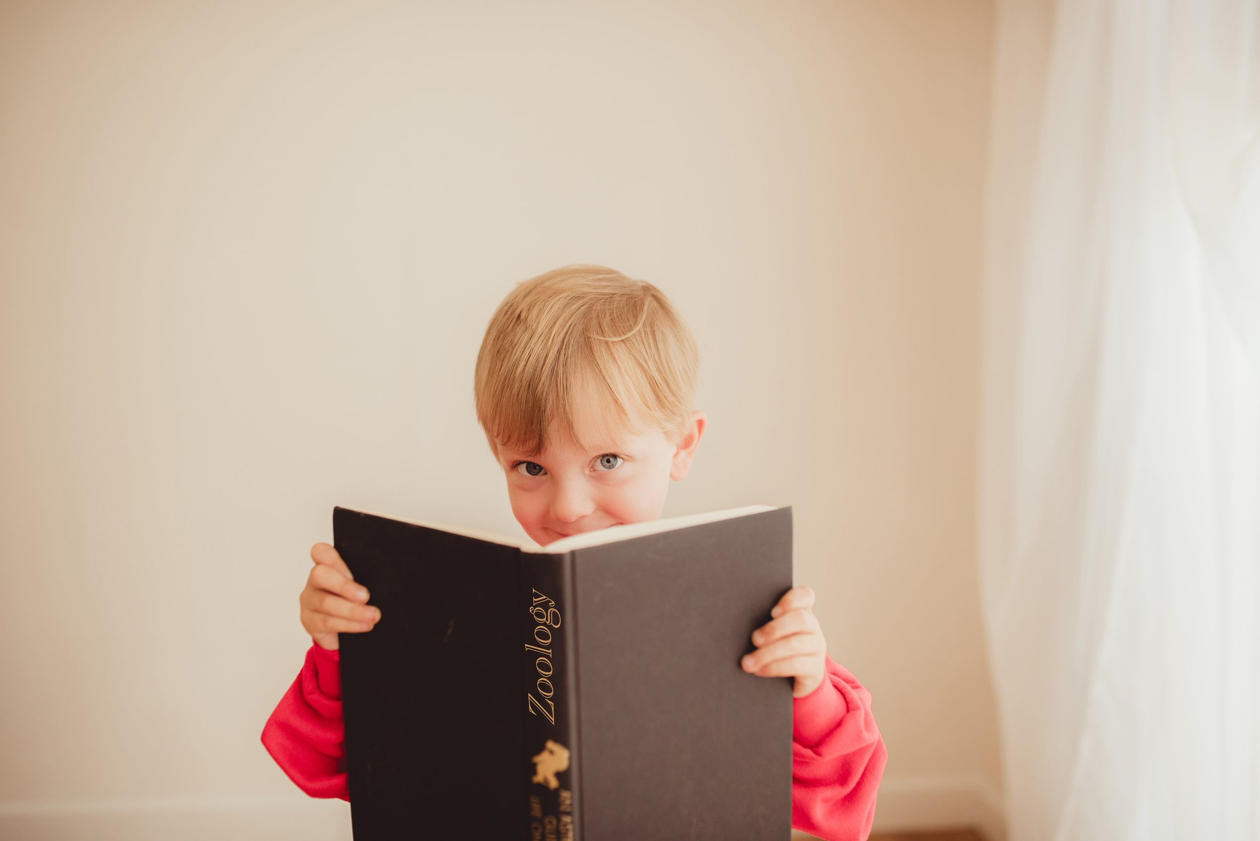 A young boy with red hair and blue eyes is wearing a red hoodie and holding a large black book titled 'Zoology'. He is peeking over the top of the book with a playful smile and looking directly at the camera, standing against a plain light-colored wa