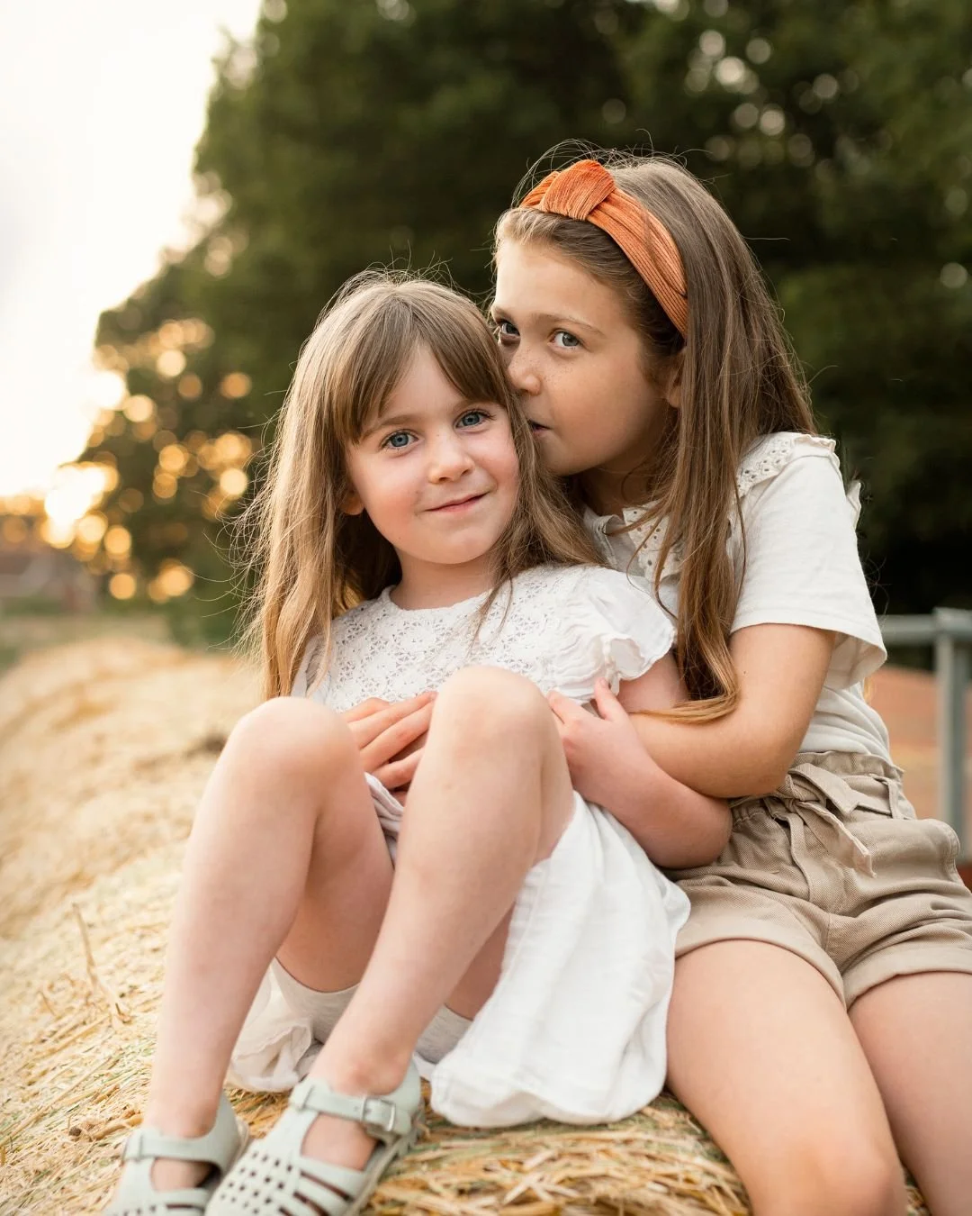Two young girls sitting on hay, one whispering to the other amidst a natural outdoor setting.