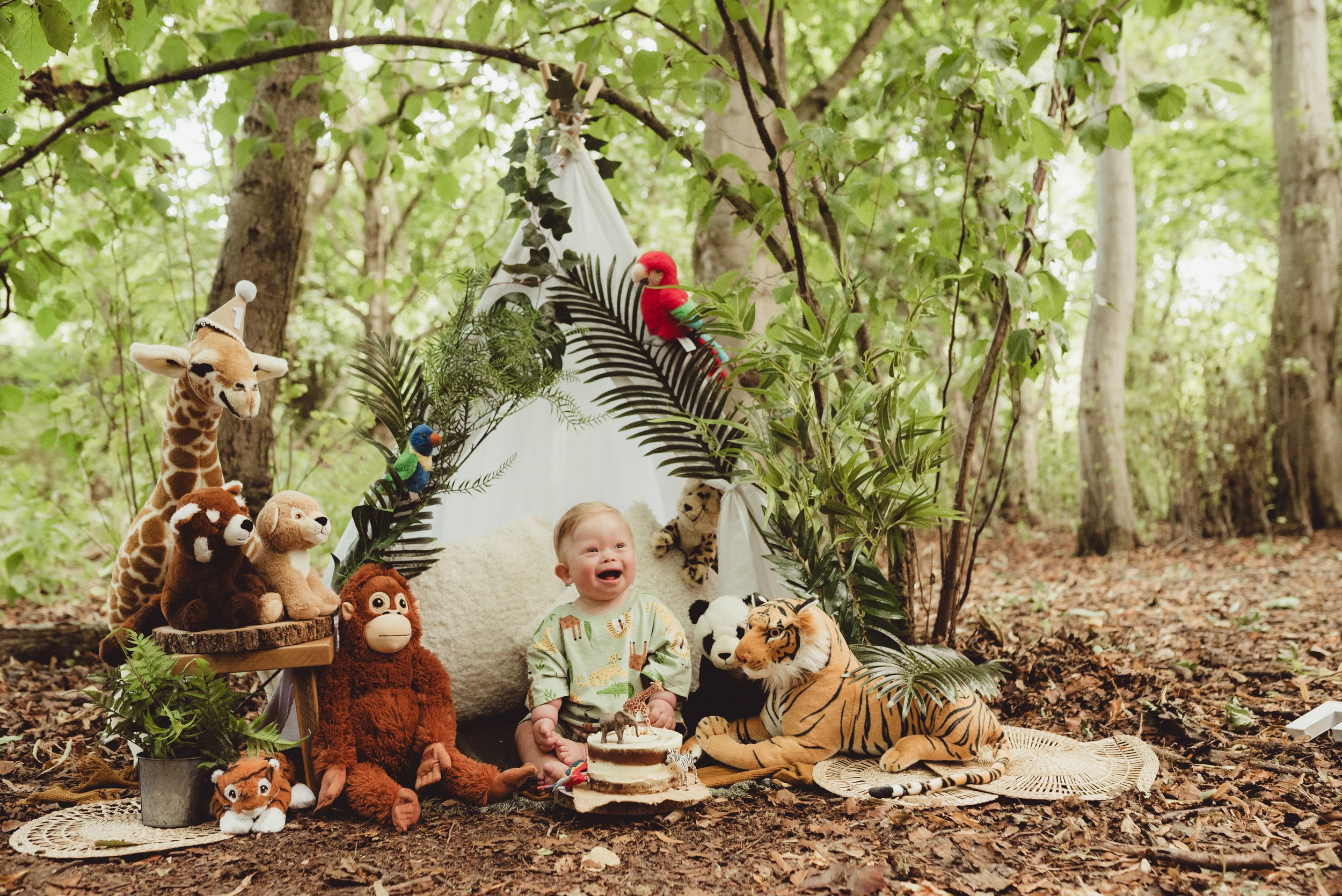 A wild one safari birthday setup in a wooded outdoor area with a small teepee decorated with greenery, surrounded by cuddly safari toys, pillows, and a cake with animal toppers.