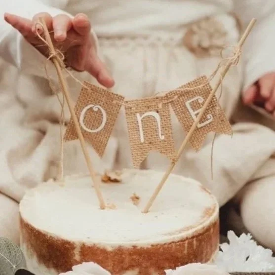 A hand holds a burlap banner spelling 'one' above a cake, with two skewers with a string of bunting attached.