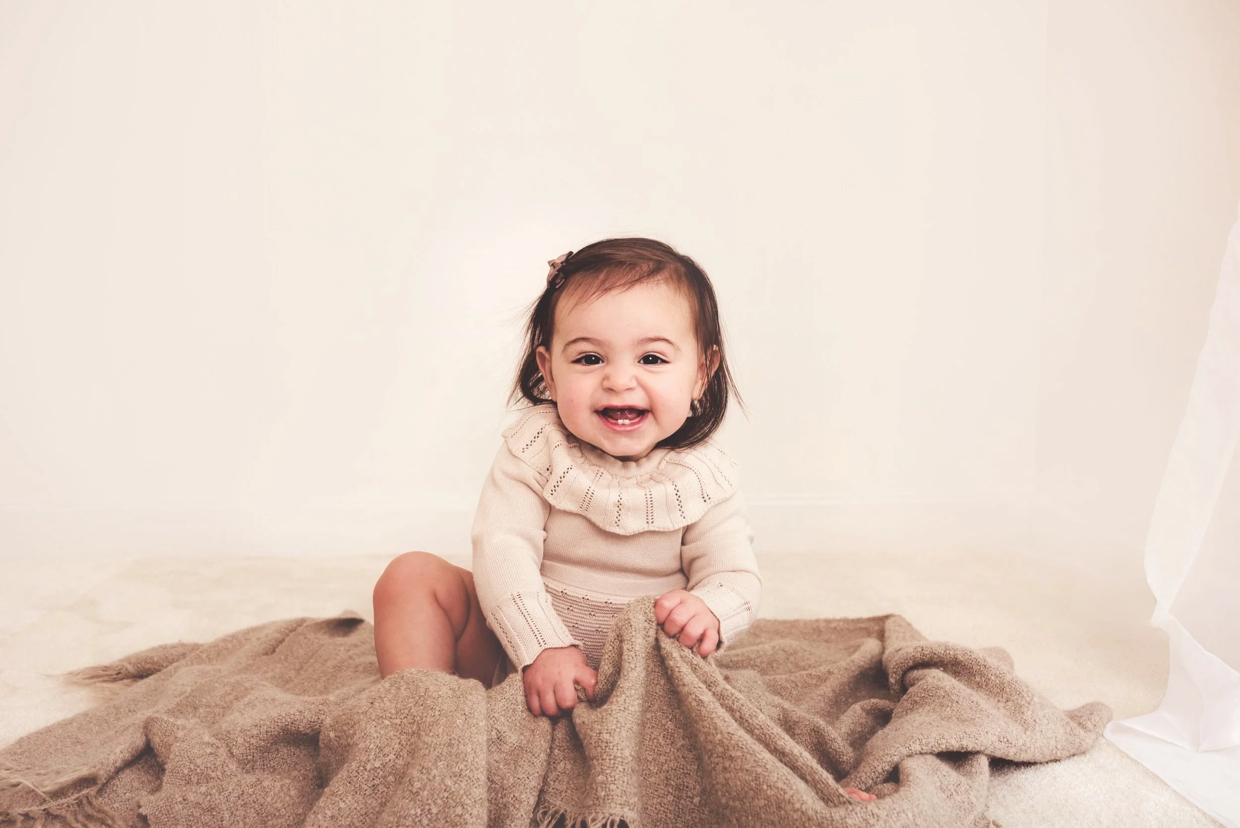 A smiling baby girl with dark hair, wearing a cream-colored sweater, sitting on a beige blanket against a plain white wall.