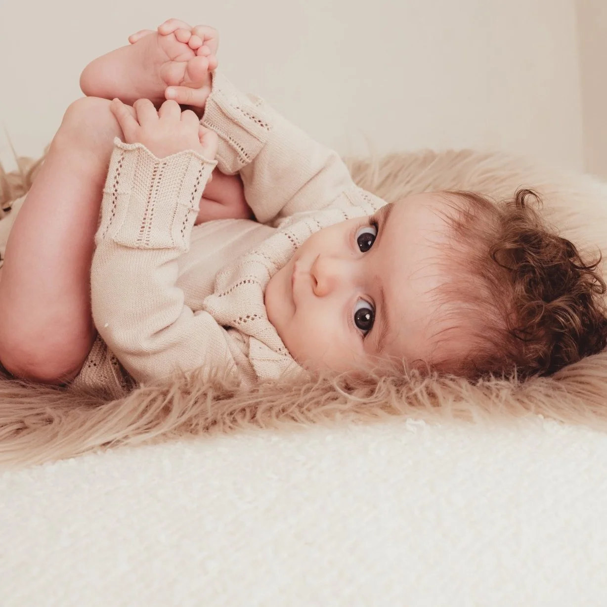A young child with curly brown hair lying on a soft, fluffy surface, holding their feet with their hands, and looking at the camera with wide eyes.