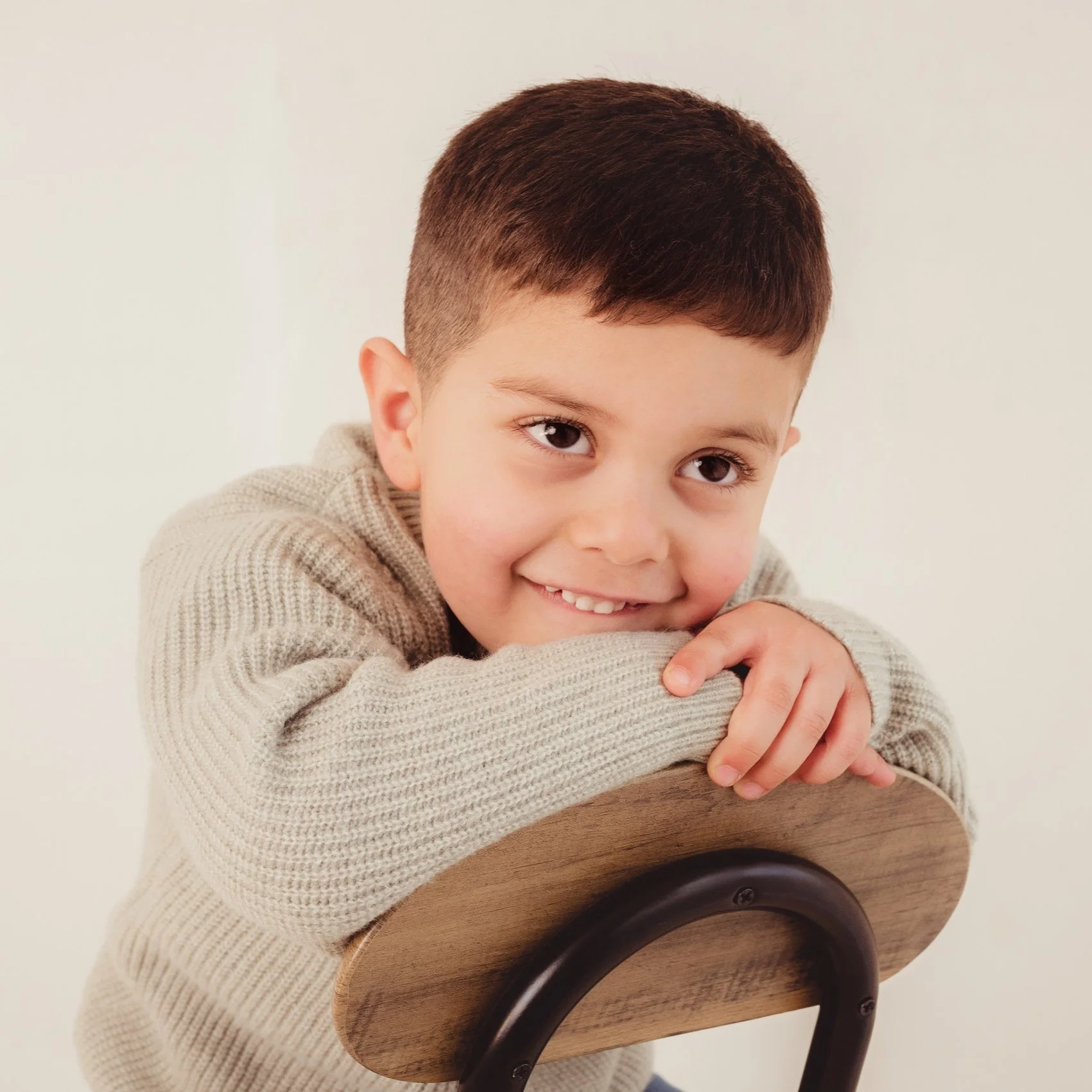 A young boy with short brown hair and a light beige sweater resting his chin on a wooden chair back, smiling at the camera.