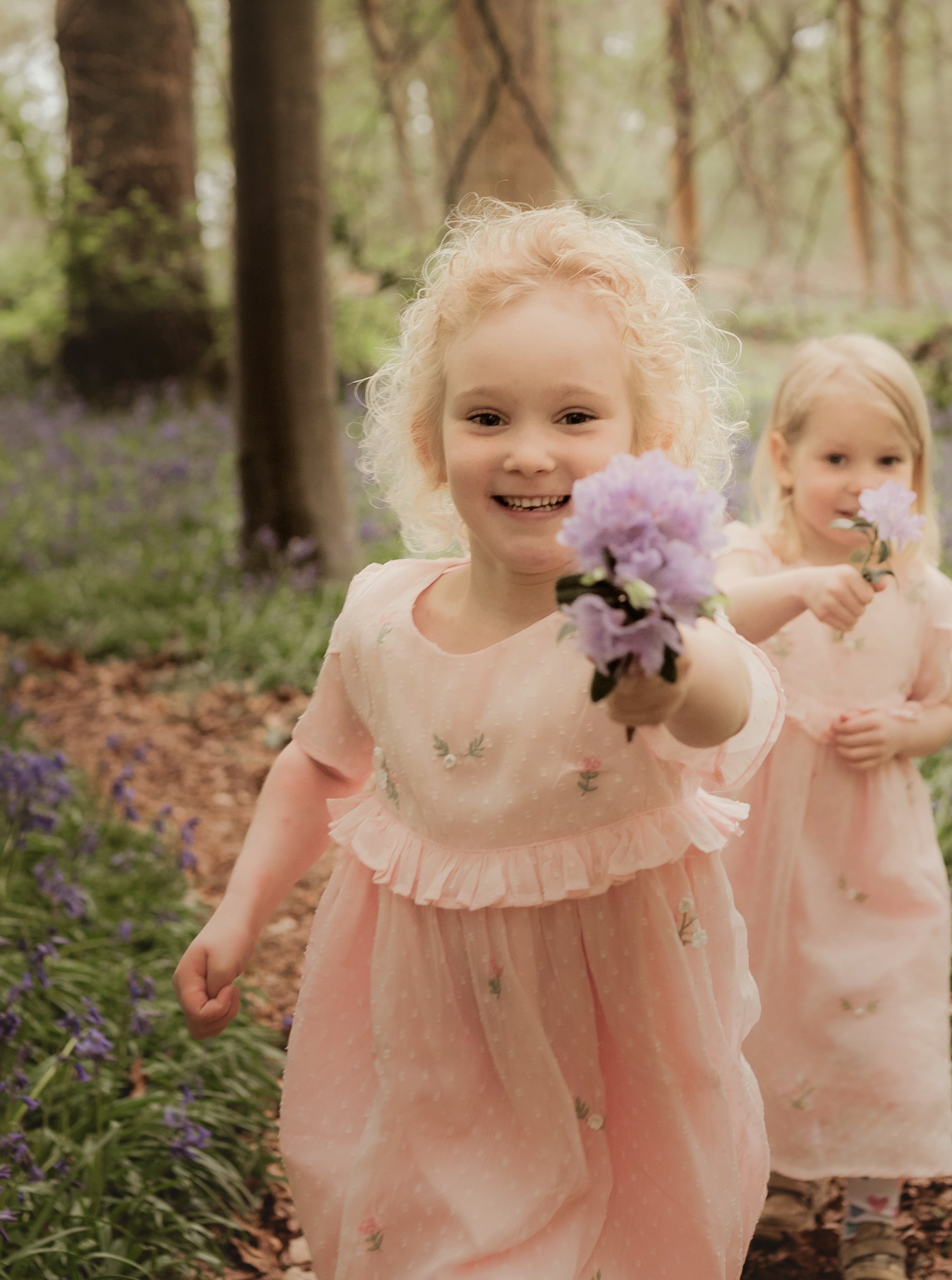 Sisters running through the woodland with bluebells in the back ground on a outdoor family photoshoot in the spring with Jodie Wilson photography, Saffron Walden Essex