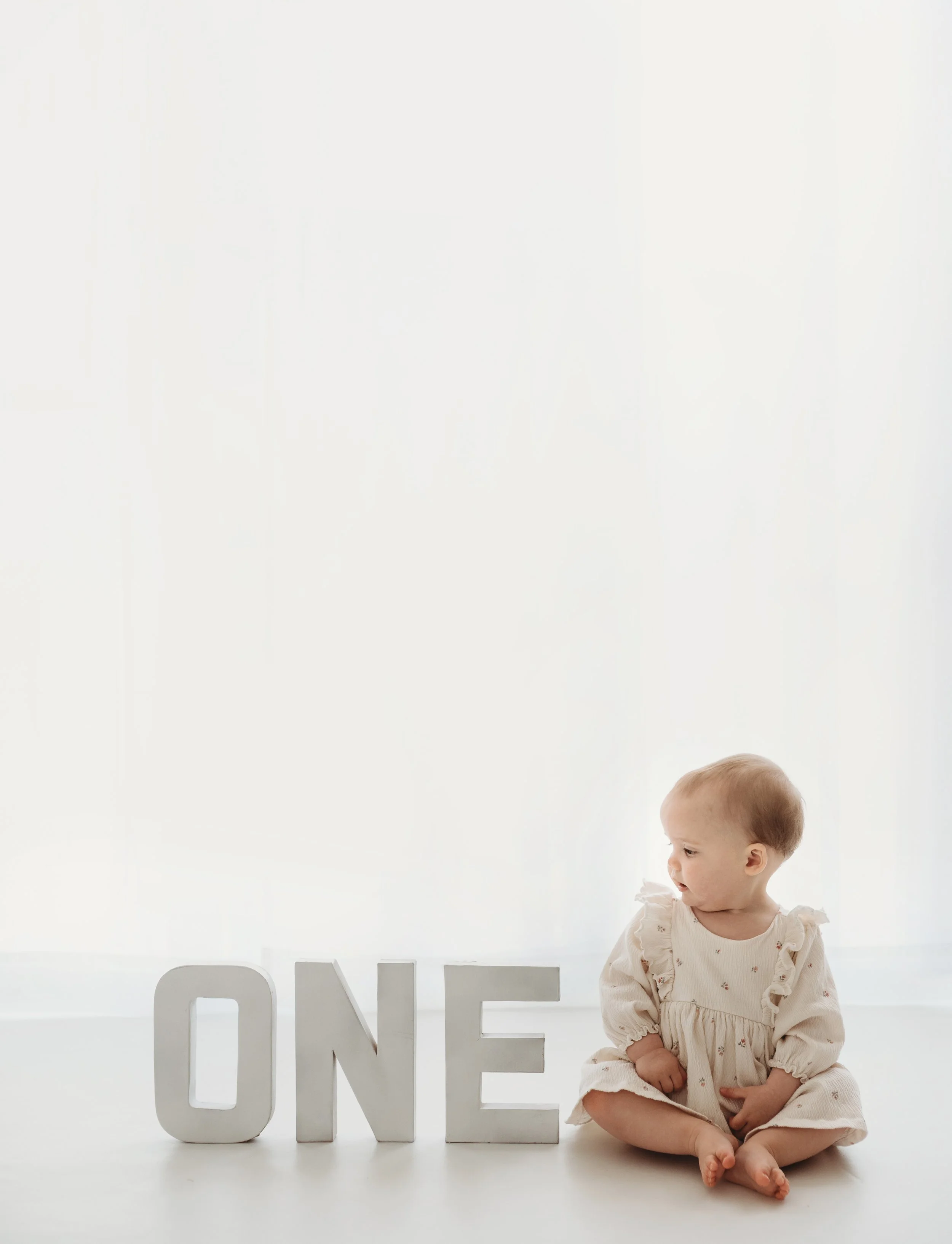A baby girl with light brown hair, wearing a cream-colored dress with ruffled sleeves, sitting on the floor next to large white decorative letters that spell 'ONE' in front of a plain white background.