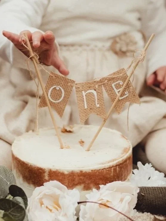 A birthday cake with a rustic style, decorated with a burlap banner that spells out 'ONE' and two wooden sticks holding the banner, surrounded by white flowers in a boho style 1st birthday cake smash.