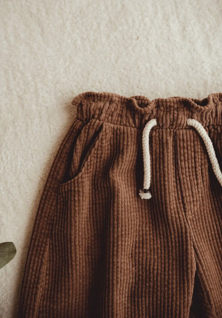 Close-up of brown corduroy pants with a white drawstring and a side pocket, hanging against a beige textured wall.