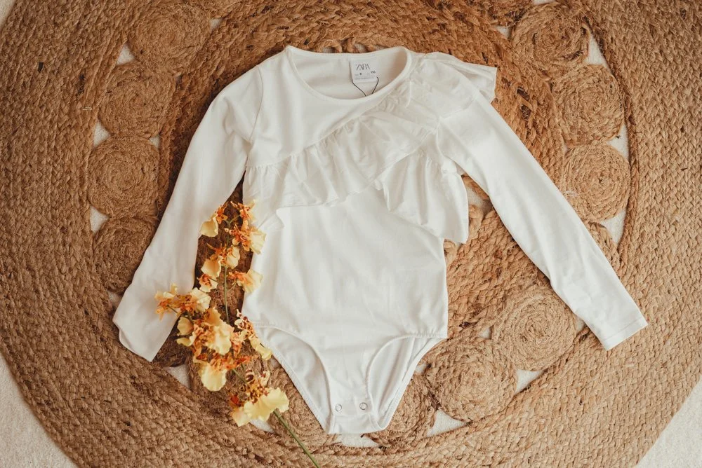 A cream-colored long sleeve bodysuit enjoying ruffle details on the chest, laid flat on a round, textured straw placemat with a bunch of peach-colored flowers beside it.