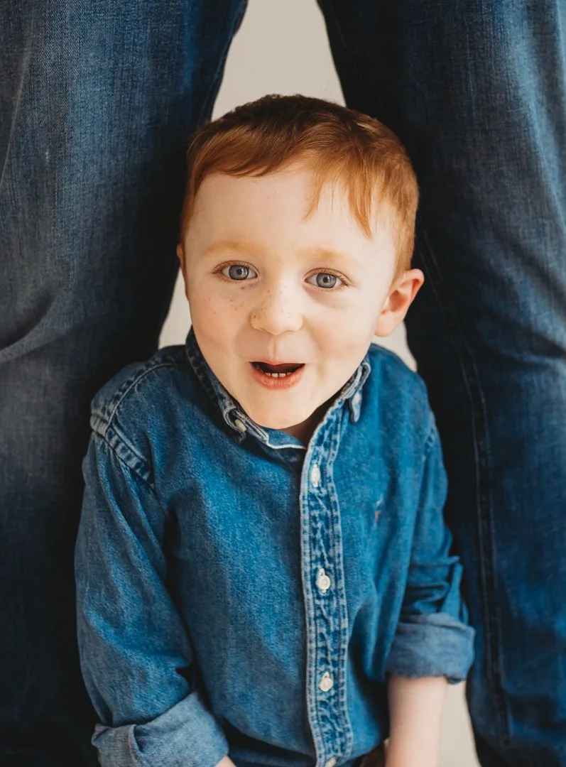 A young boy with red hair and blue eyes, wearing a denim shirt, looking up and smiling, with an adult in blue jeans behind him.