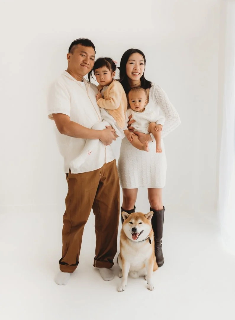 Family portrait with two adults, two young children, and a dog standing on a white background.