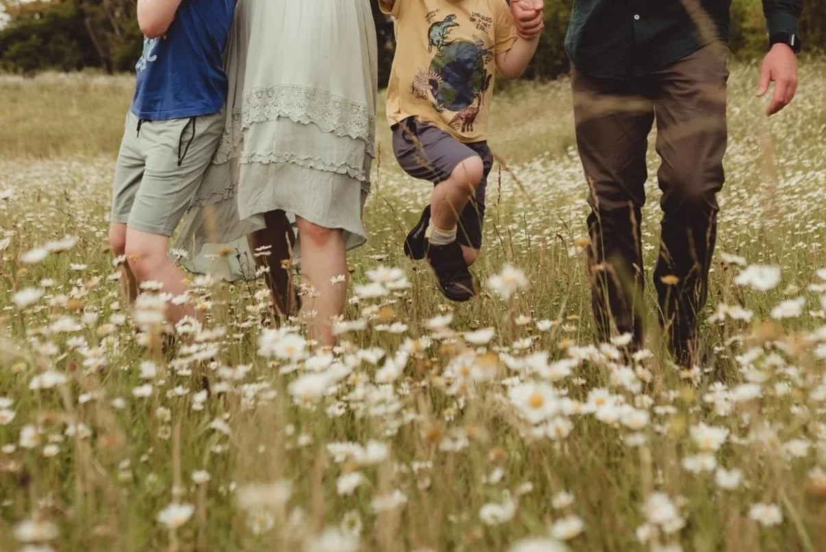 A family of three walking through a field of daisies, showing the legs and lower bodies of an adult woman, a young boy, and an adult man.