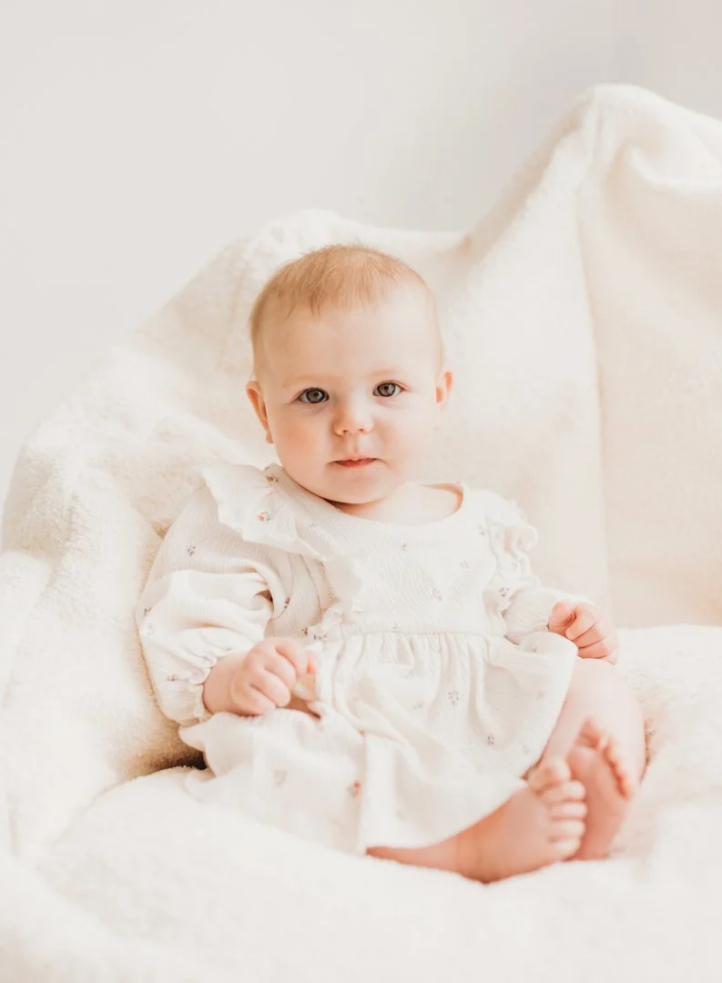 A baby girl with red hair and blue eyes sitting on a soft white blanket, wearing a cream-colored dress with ruffles.
