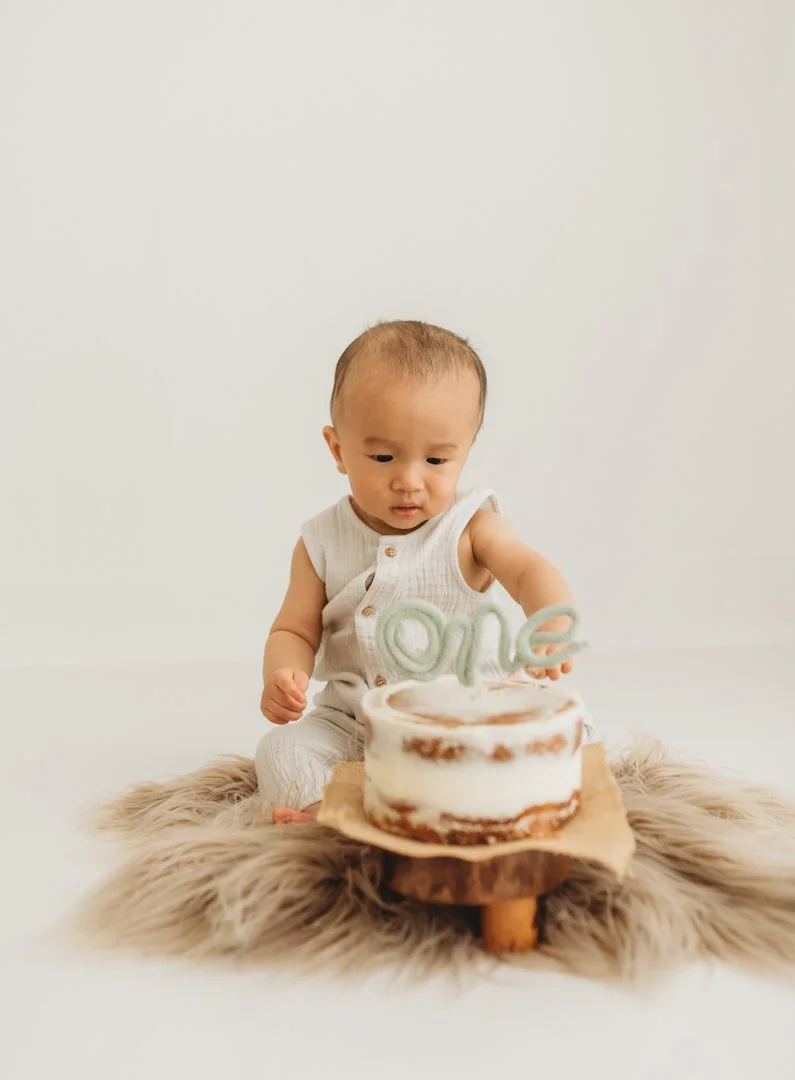 A young child sitting on a faux fur rug, looking at a small birthday cake with a 'one' cake topper.