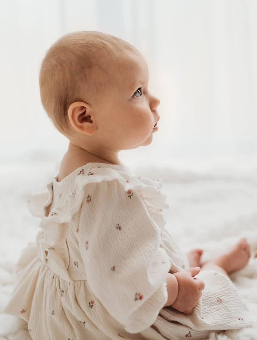 A young child sitting on a white surface, looking to the side. The child has light hair, fair skin, and is wearing a cream-colored dress with small floral patterns.
