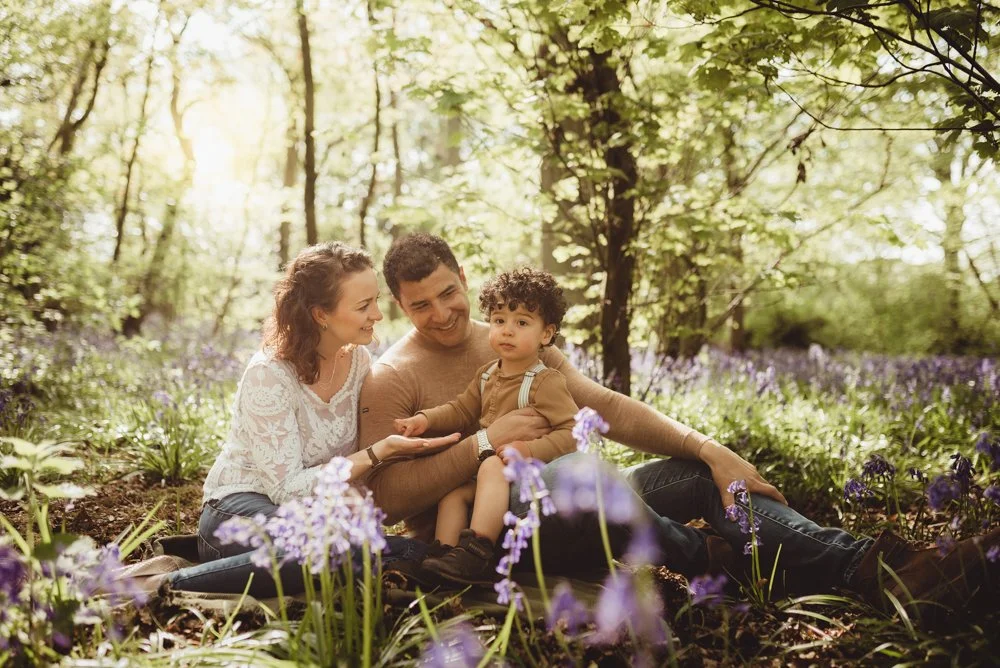 A family of three, consisting of a woman, a man, and a young boy, sitting on the ground in a wooded area with purple flowers, enjoying a sunny day.