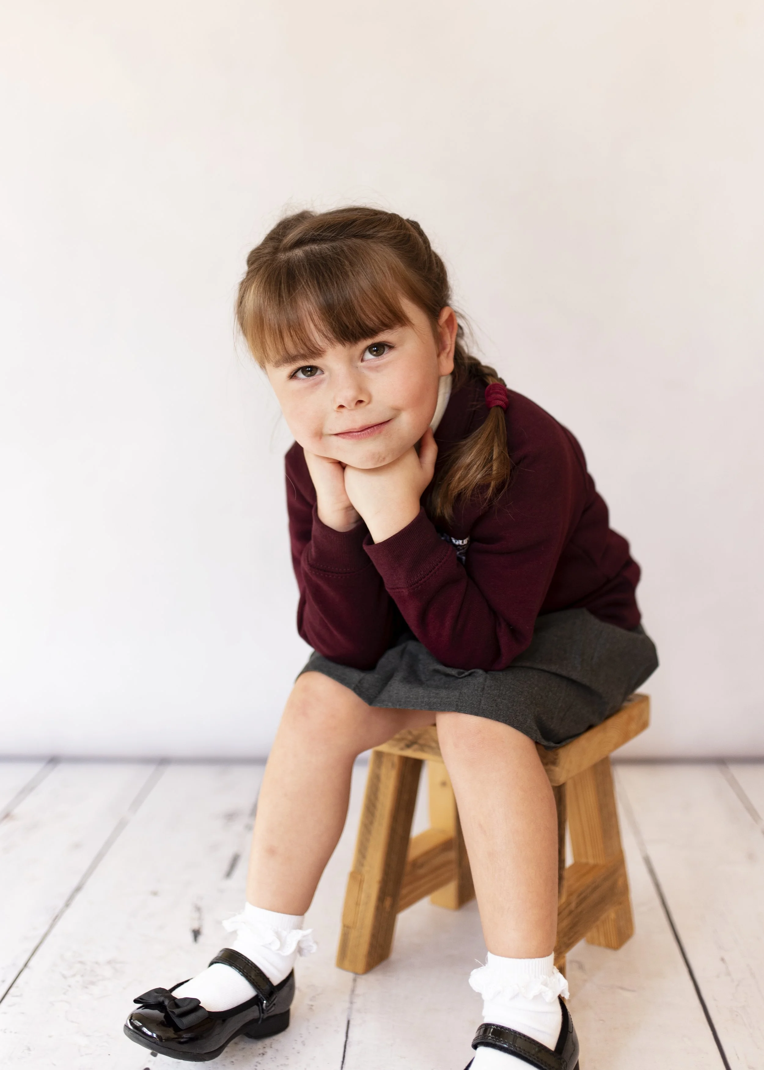 A young girl with brown hair and bangs, wearing a maroon sweater and a gray skirt, sits on a wooden stool against a plain white wall. She has her chin resting on her hand and is smiling slightly, with one leg crossed over the other. She is wearing wh