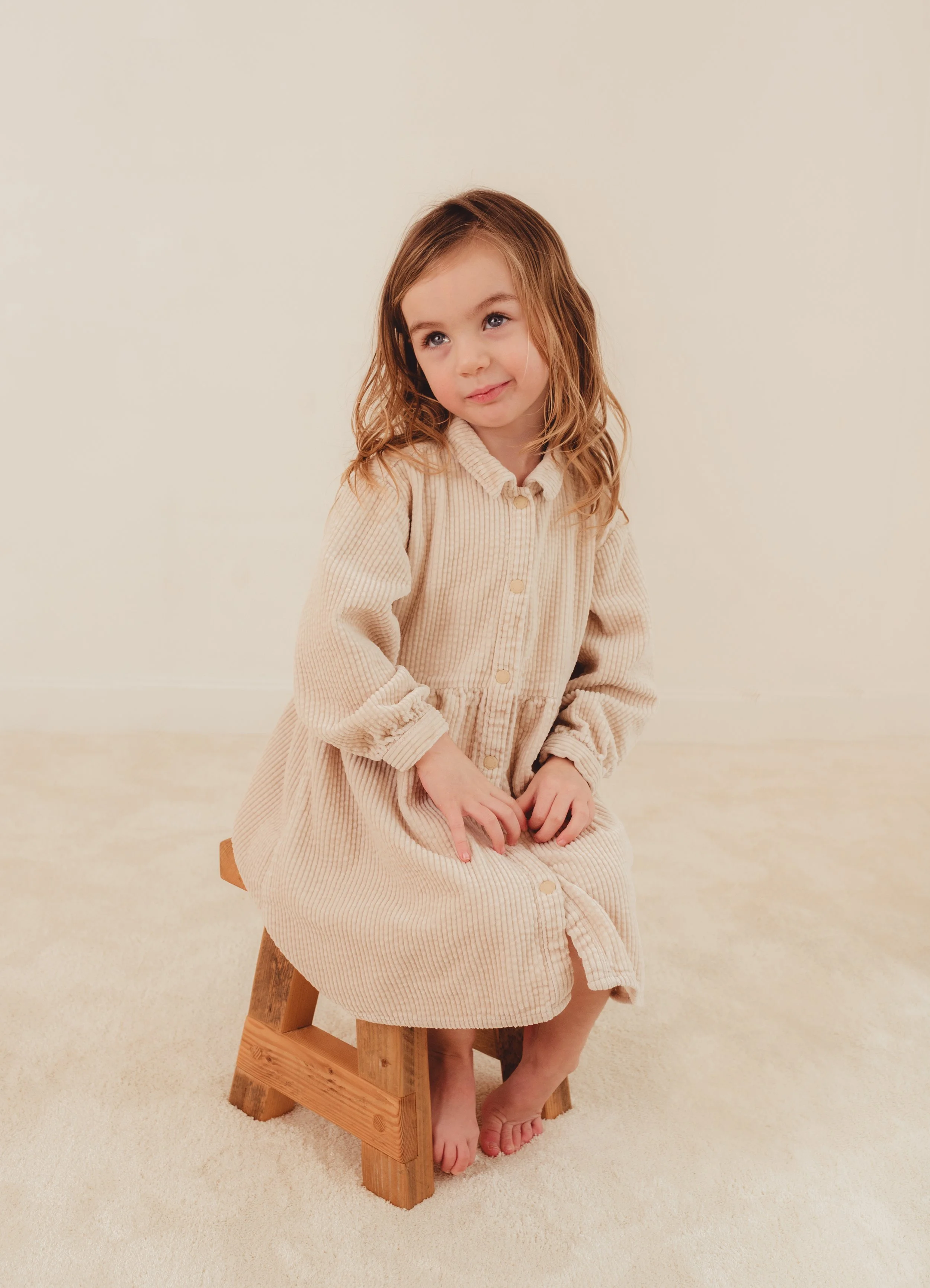 A young girl with wavy, light brown hair and blue eyes, wearing a beige, long-sleeve, button-up dress with a collar, sitting barefoot on a small wooden stool in a plain, light-colored room.