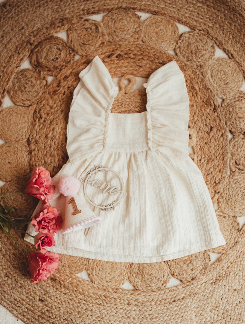 A cream-colored toddler dress with ruffled straps laid on a round woven rug. Next to the dress are pink carnations, a small pink hat with a pom-pom, a silver ring with the word 'Love,' and a white ring with a number one, indicating a first birthday c