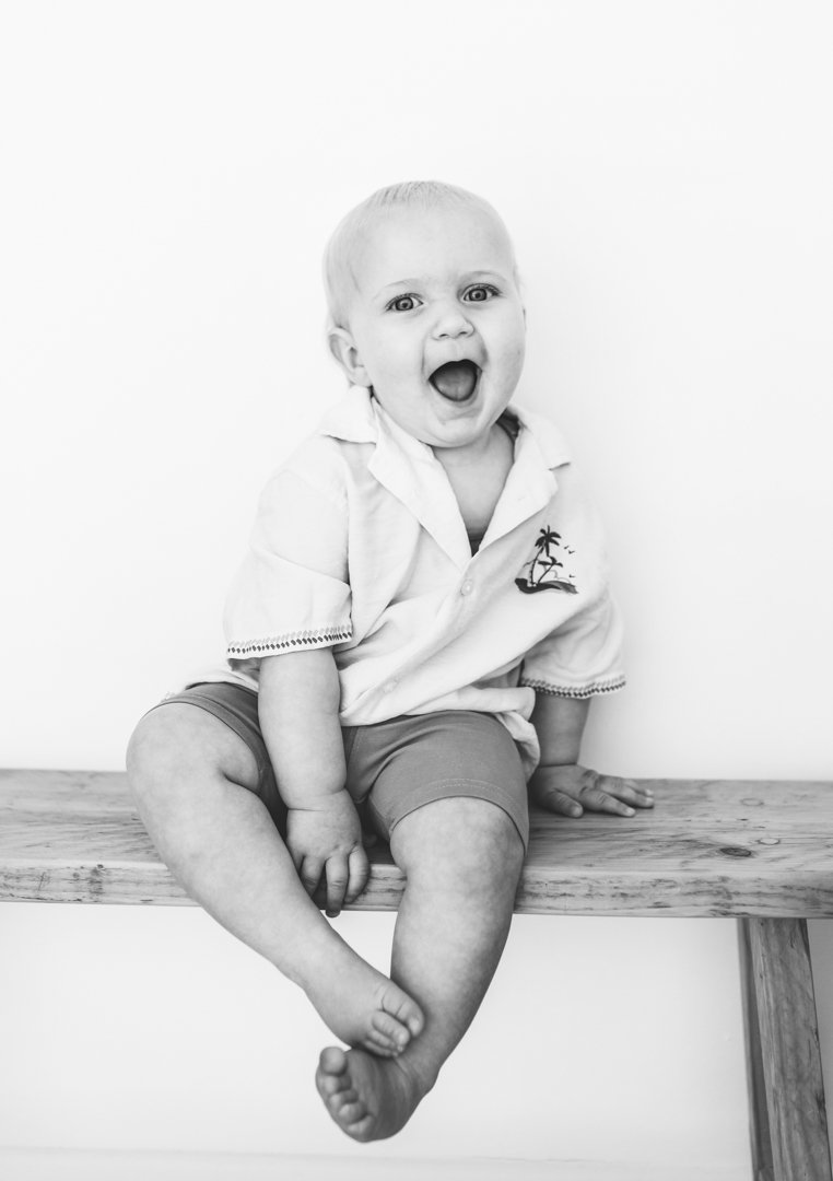 Black-and-white photo of a toddler sitting on a wooden bench, looking surprised or excited, with a wide-open mouth and big eyes, wearing a collared shirt with a palm tree design and shorts.