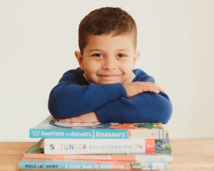 A young boy with short brown hair, wearing a blue sweater, resting his arms on a stack of six colorful children's books about dinosaurs.