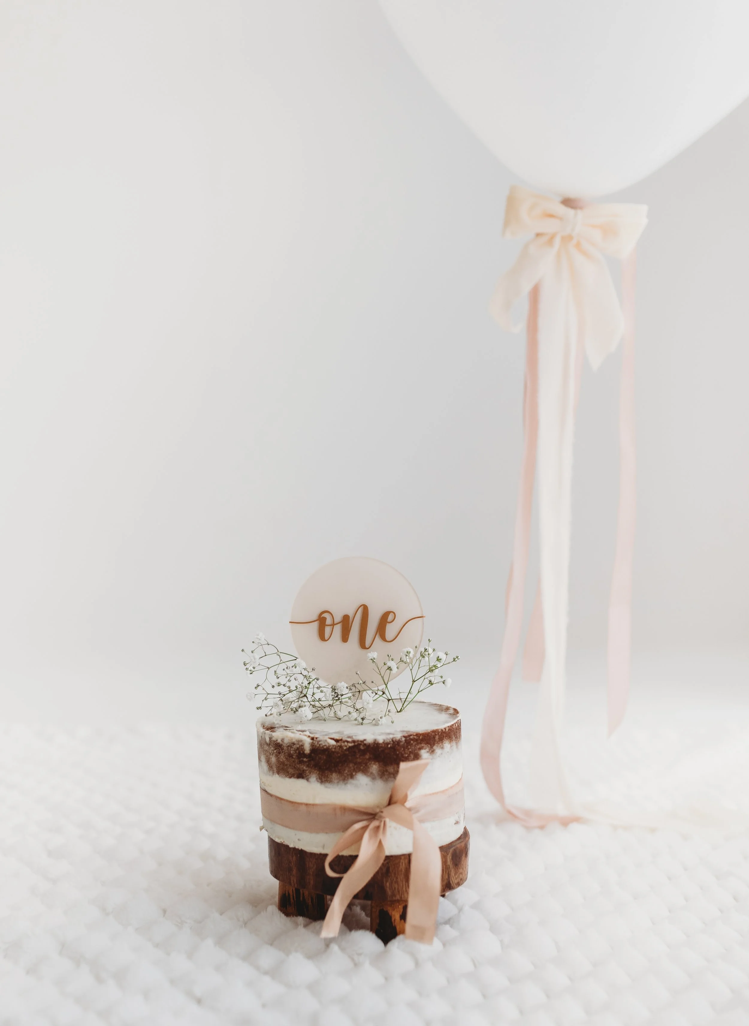 A small wooden log decorated with a pink ribbon bow, white frosting, dried baby's breath flowers, a round topper with the word "one" in cursive, a pink and white balloon, and ribbons, set against a white background for a first birthday celebration.
