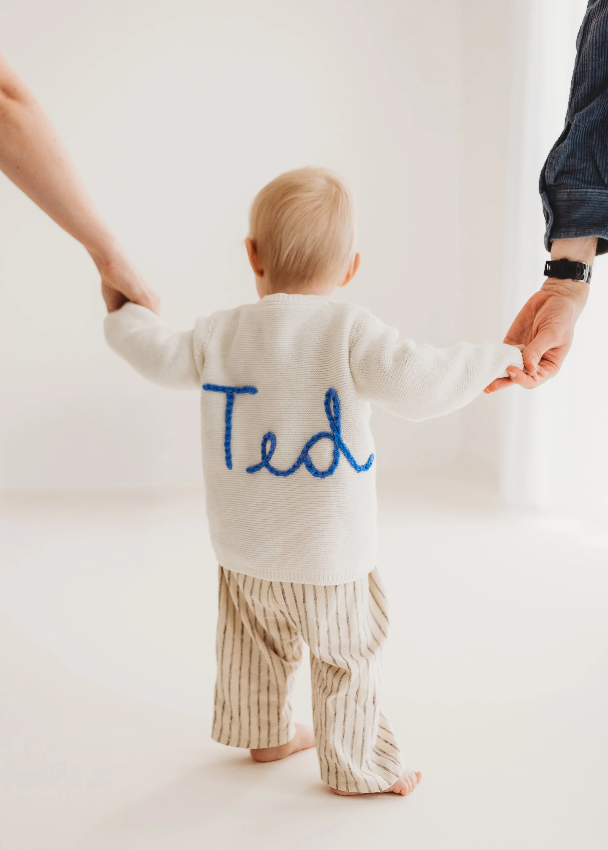 Back view of a toddler with blonde hair walking barefoot, holding hands with two adults, one on each side, in a neutral room. The child wears a personalised knitted jumper with the word 'Ted' embroidered in blue and striped beige pants.