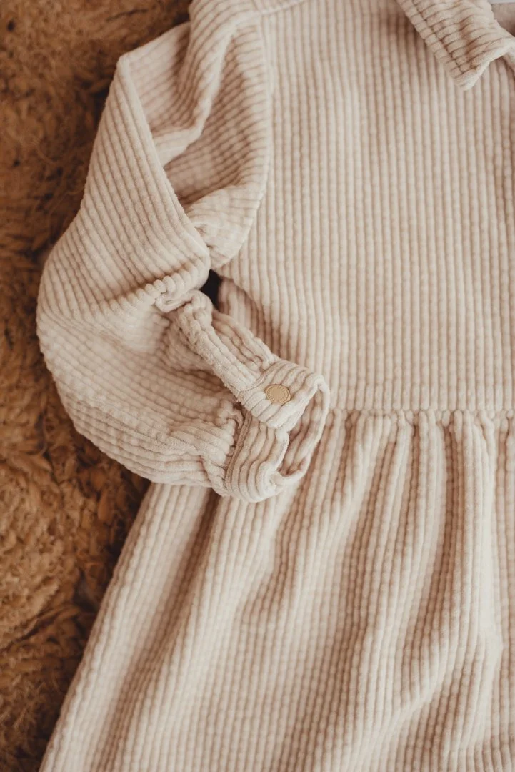Close-up of a cream-colored, ribbed, long-sleeve dress with gathered waist and rolled-up sleeve secured with a button, lying on a textured brown surface.