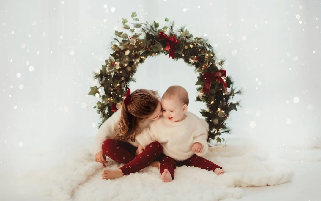 Siblings sitting on a fluffy white surface under a Christmas wreath decorated with red ribbons, holly, and lights, sharing a joyful moment.