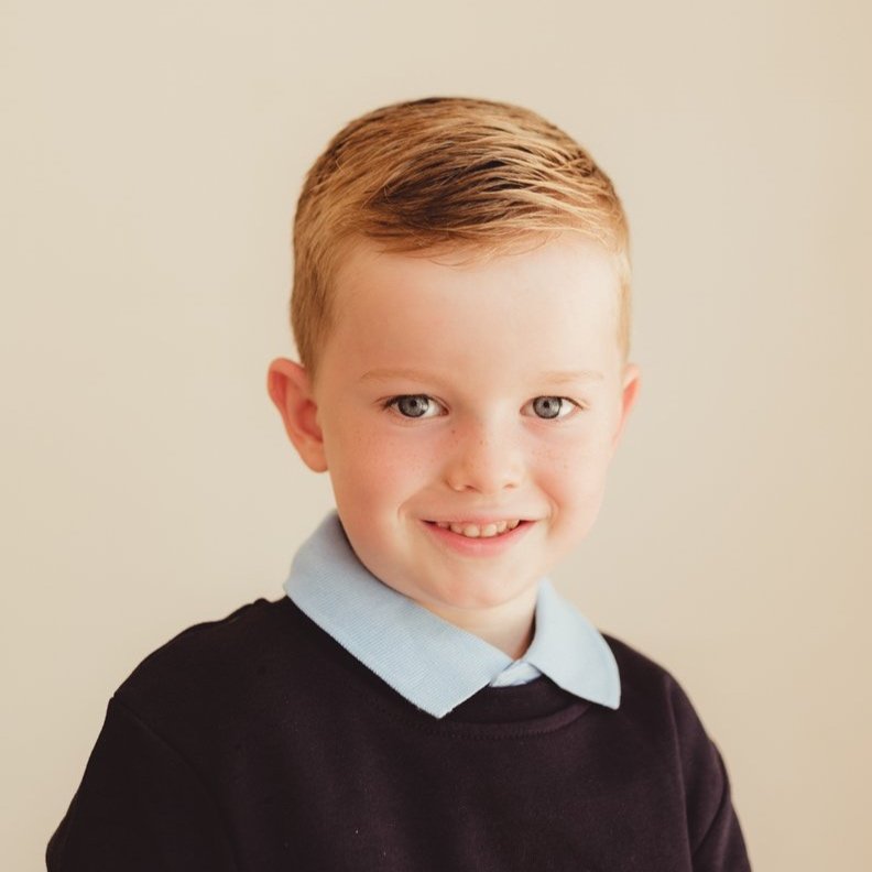 A young boy with short red hair, blue eyes, and fair skin smiling, wearing a light blue collared shirt underneath a black sweater, against a plain light-colored background.